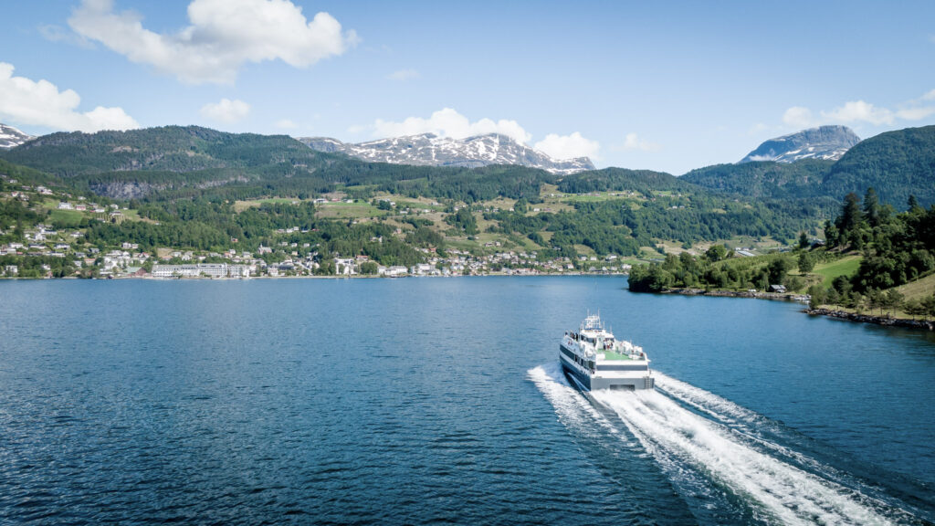 A passenger boat sailing into Ulvik in Hardanger. Green fields and forest, snow patches on mountains behind.