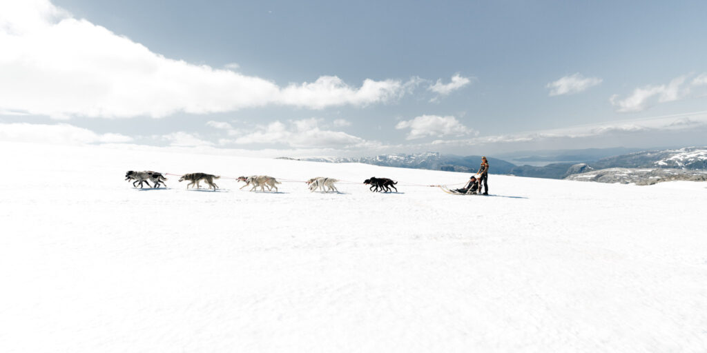 Dogsledding across a snowy mountain with dogs pulling a sled with wto people.