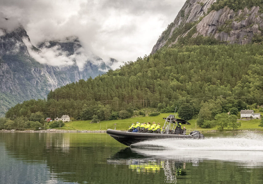 A Rib boat with passengers in yellow floatation suits speeding on a fjord with mountains covered with green trees in the background.