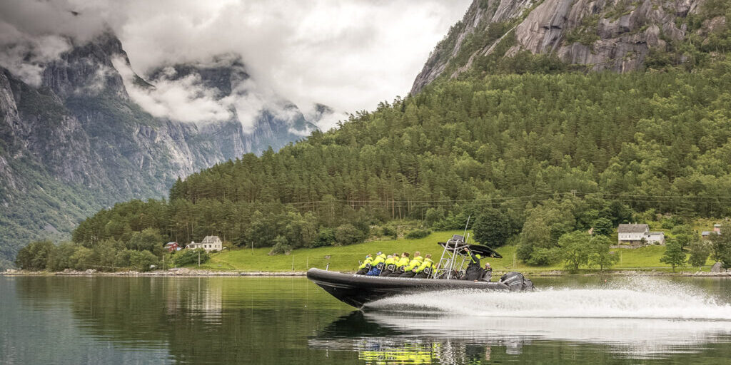 A Rib boat with passengers in yellow floatation suits speeding on a fjord with mountains covered with green trees in the background.