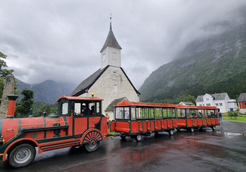 Red and green mini train on wheels (the Trolltrain) parked in front of Eidfjord church.