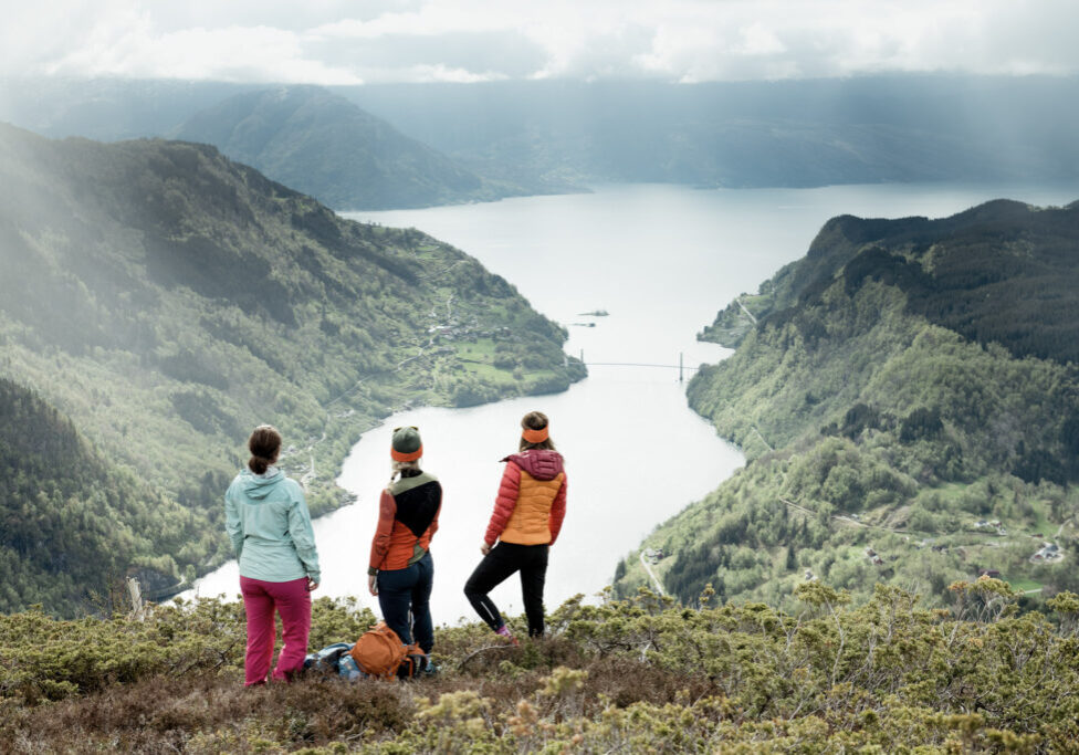 Three woman on a mountain slope looking down at the fjord and bridge below them. 