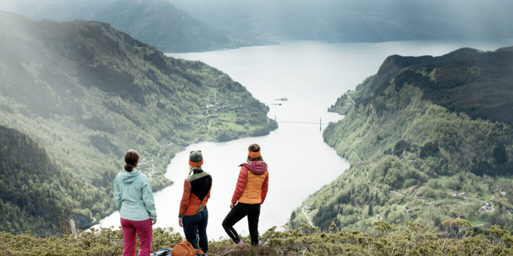 Three woman on a mountain slope looking down at the fjord and bridge below them. 