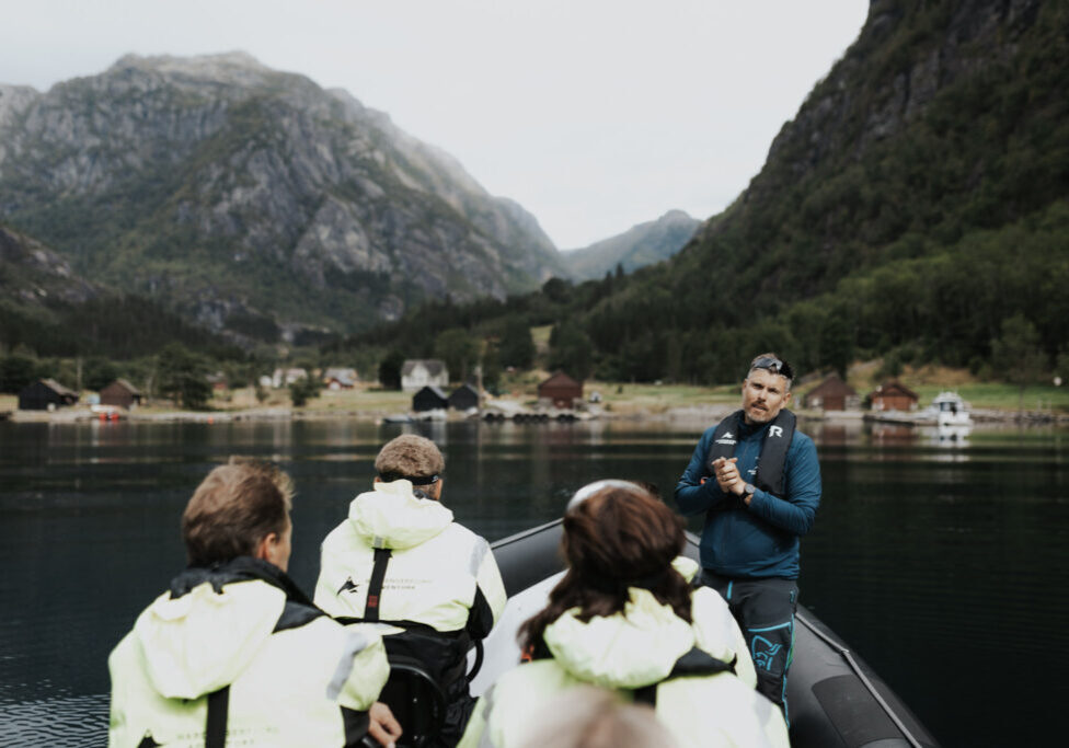 Three passengers in floatation suits listening to their guide on a RIB boat parked by fjord shore.