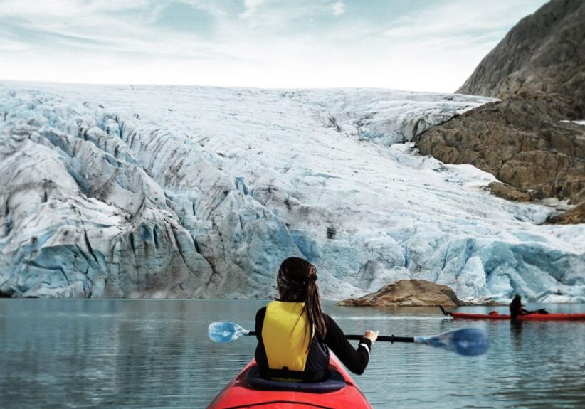 A person in a red kayak seen from the back, paddling in front of a glacier with another kayak in the background.