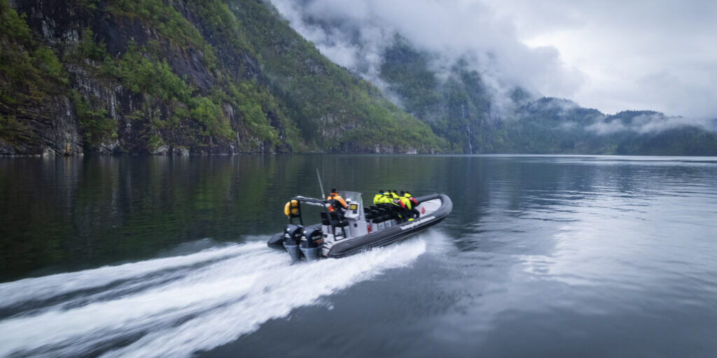 RIB boat with passengers in motion along a fjord shoreline on a cloudy day.