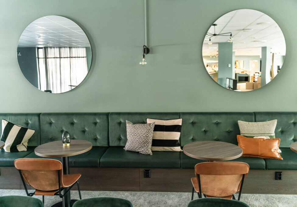Green leather benches in front of a green wall with two sircular mirrors. Round tables and cognac coloured leather chairs.