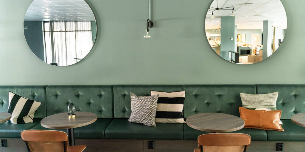 Green leather benches in front of a green wall with two sircular mirrors. Round tables and cognac coloured leather chairs.