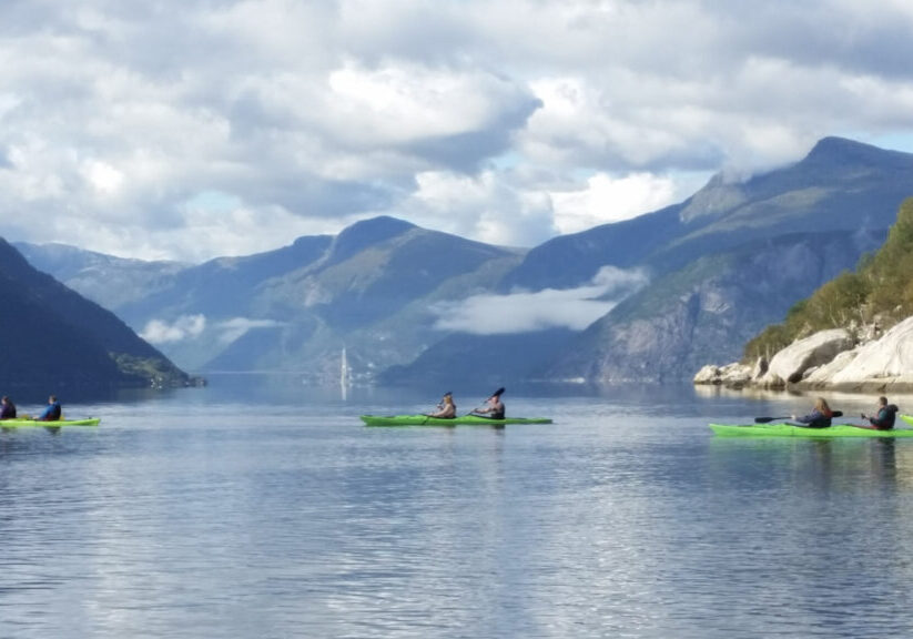 Four green double kayaks on a fjord with mountains and a bridge in the background.