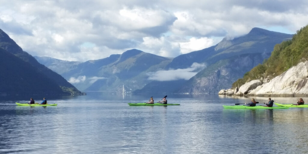 Four green double kayaks on a fjord with mountains and a bridge in the background.