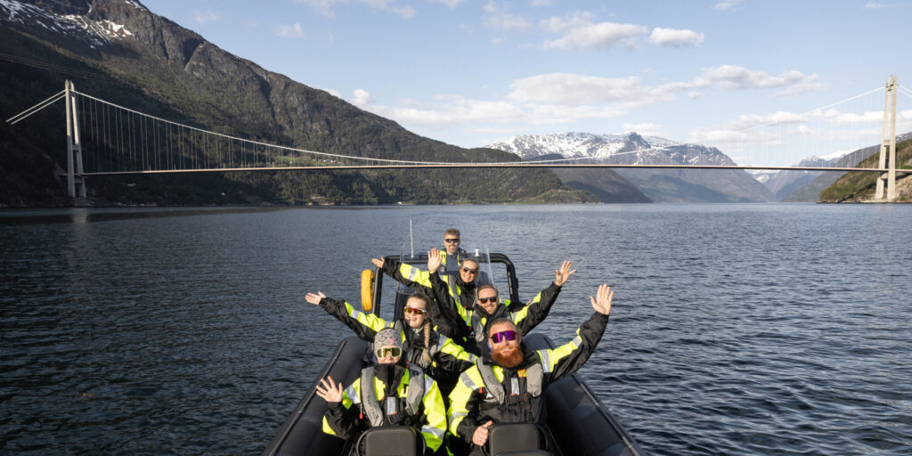Happy people in floatation suits on a RIB boat on a fjord with Hardanger bridge in the background.