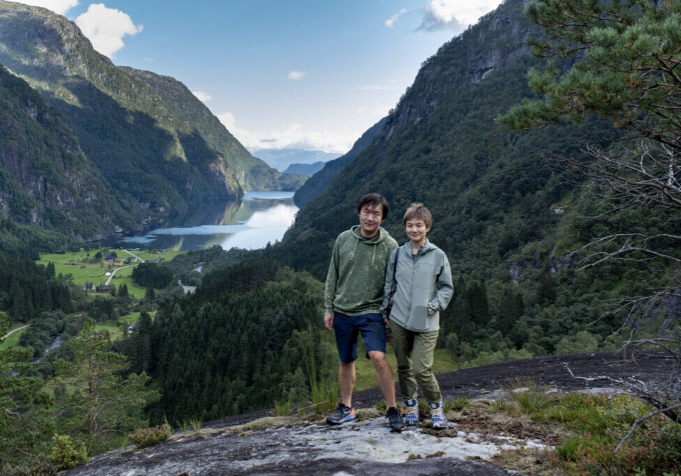 Two people posing on top of a mountain with a fjord in the background.