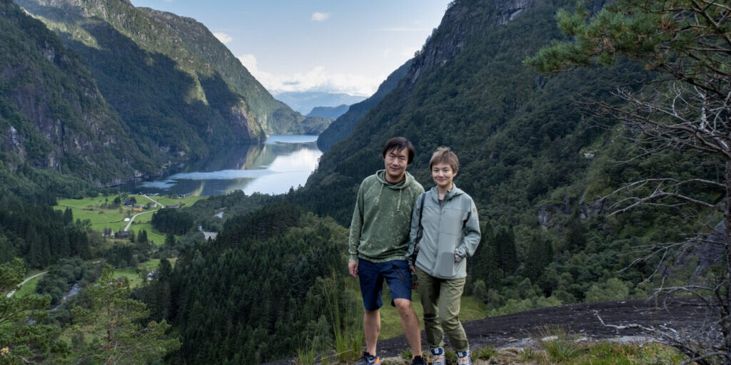 Two people posing on top of a mountain with a fjord in the background. 