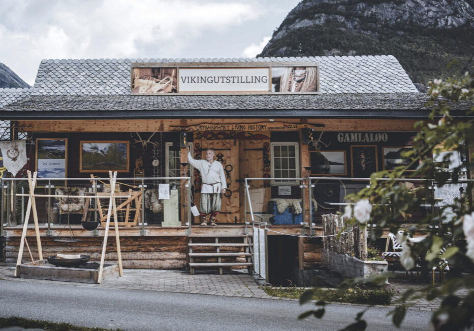 A person dressed like a Viking standing in front of a wooden house with "Vikingutstilling" sign.