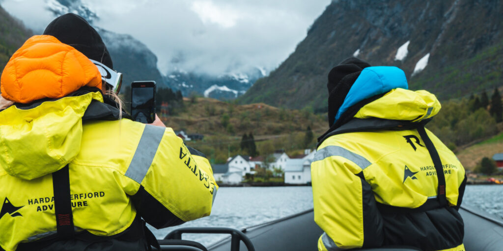 Two people in yellow floatation suits on a boat, one of whom is taking a picture with a mobile phone.