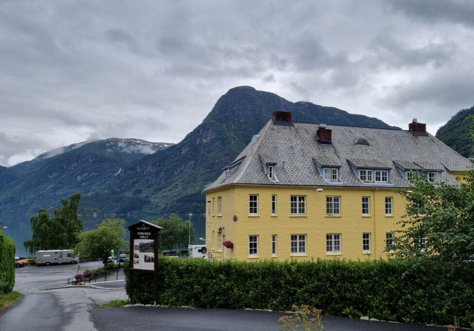 Yellow hotel building with grey roof surrounded by green bushes, with mountains in the background.